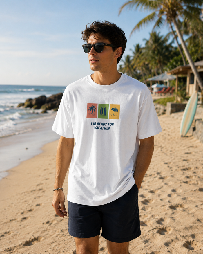 A man wearing a white graphic t-shirt and dark shorts stands on a sandy beach with palm trees and the ocean in the background. The t-shirt features a colorful graphic of three beach-themed icons above the text 'I'M READY FOR VACATION'.