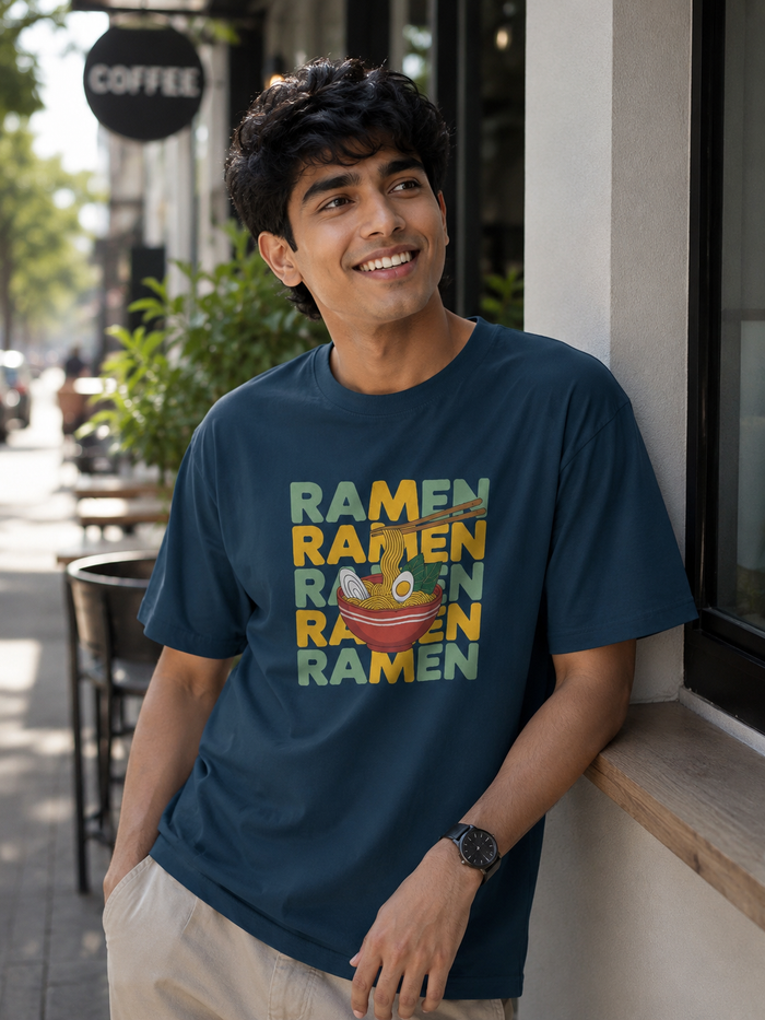 A man smiling and leaning against a storefront window wearing a navy blue t-shirt with a colorful graphic that reads 'RAMEN RAMEN RAMEN RAMEN' above and below a bowl of ramen.