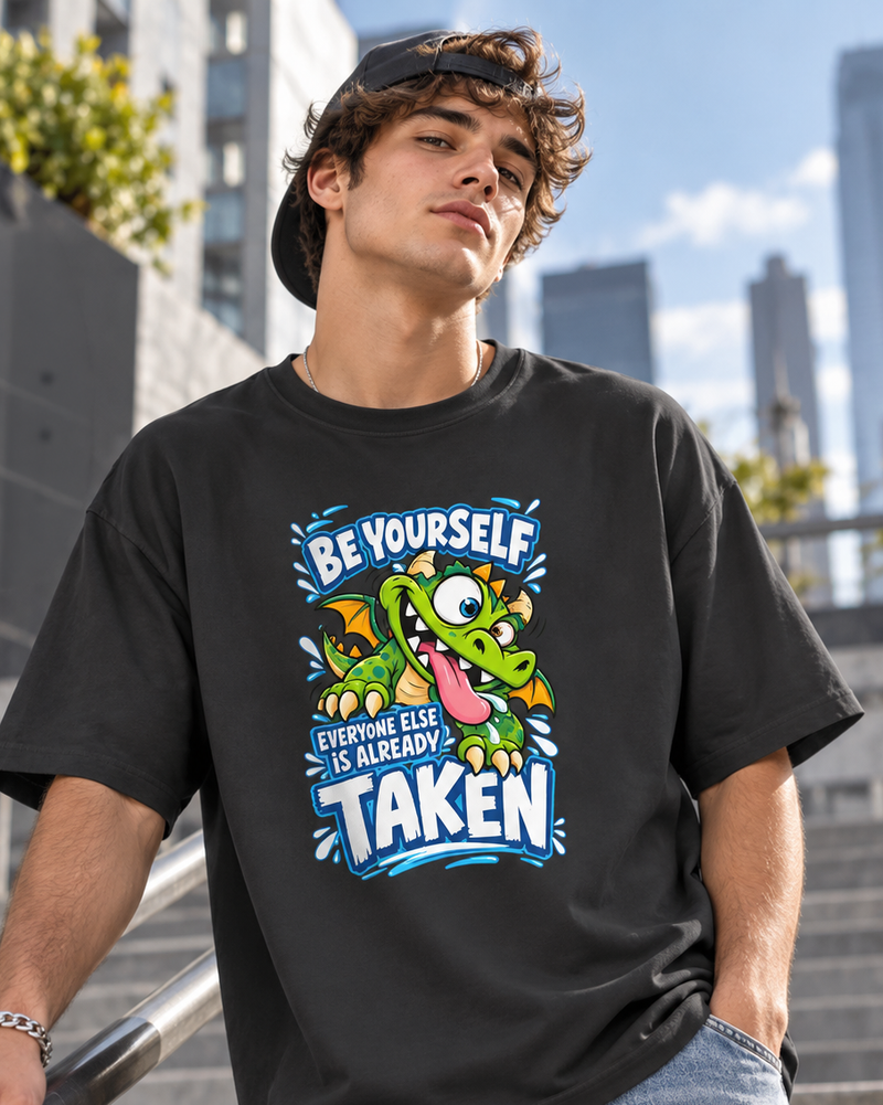 A man wearing a black graphic t-shirt with a cartoon green dinosaur and the text 'BE YOURSELF EVERYONE ELSE IS ALREADY TAKEN'. The background shows an outdoor urban setting with blurred skyscrapers and stairs.